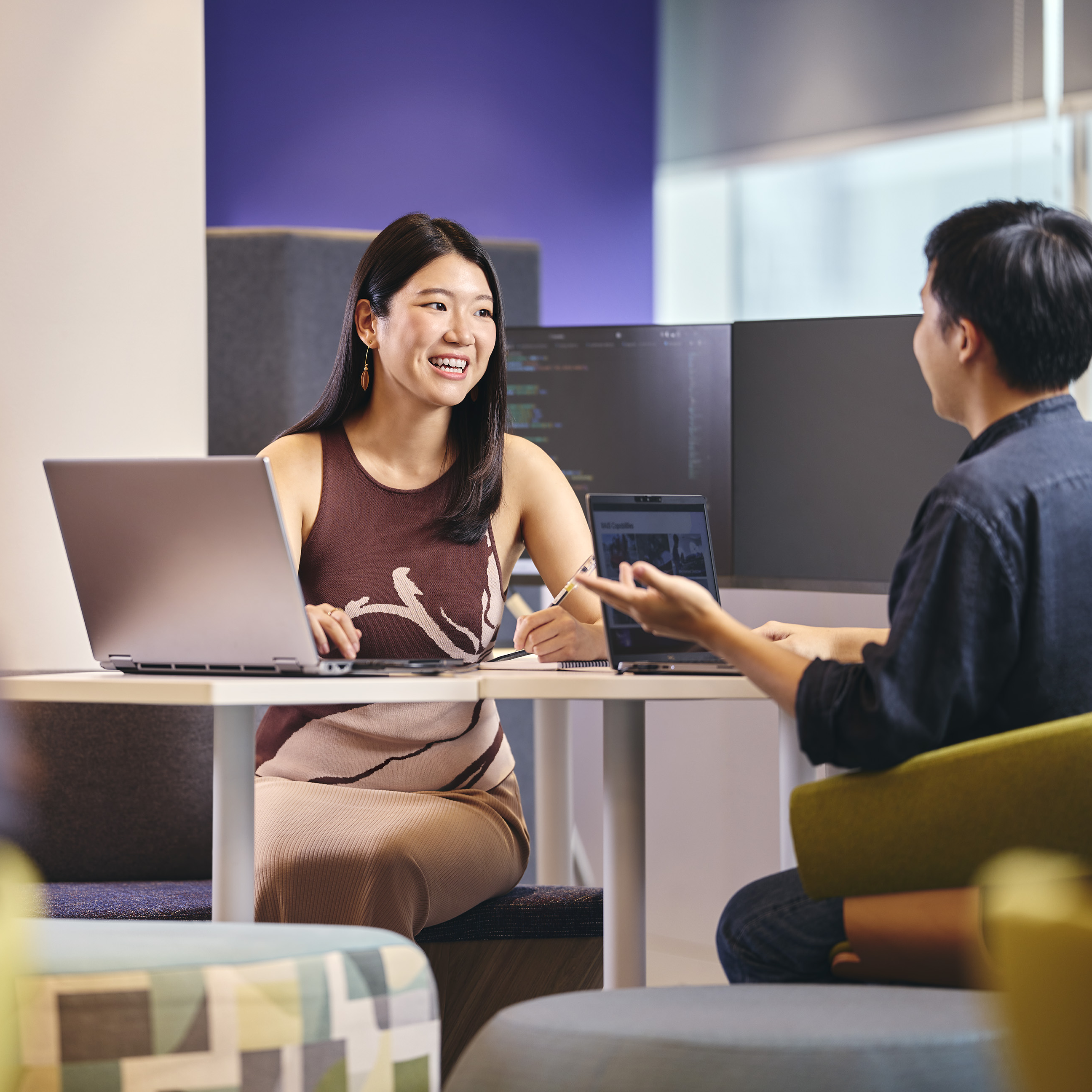 Two people at a table with laptops and monitors in an office setting.
